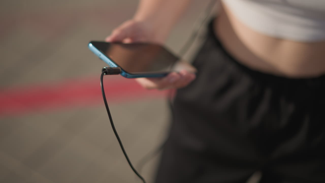young white dancer on street preparing playlist, holding smartphone and wired headphones, scrolling tracks and adjusting cord, revealing midriff in white crop top and black jogger pants, late