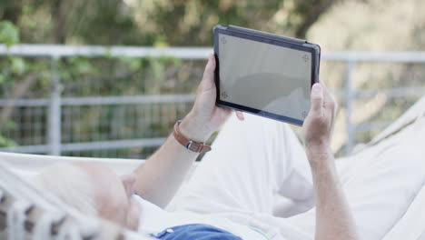 Kaukasischer-Mann-Mittleren-Alters,-Der-Ein-Tablet-Benutzt-Und-In-Der-Hängematte-Auf-Der-Terrasse-In-Der-Natur-Entspannt,-Zeitlupe