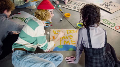 Back-view-of-young-environmental-activists-painting-placards-sitting-on-the-floor