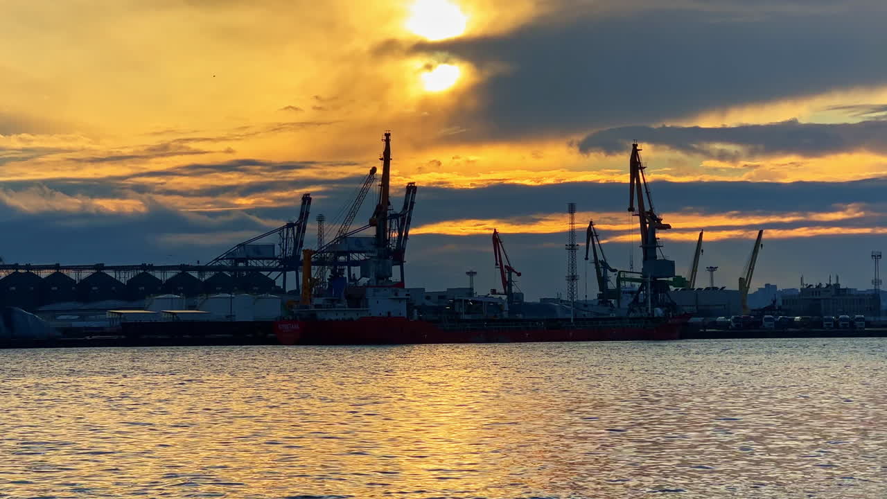 Solnedgång över hamnen med kranar och fartyg vid en livlig hamn under kvällstid.
