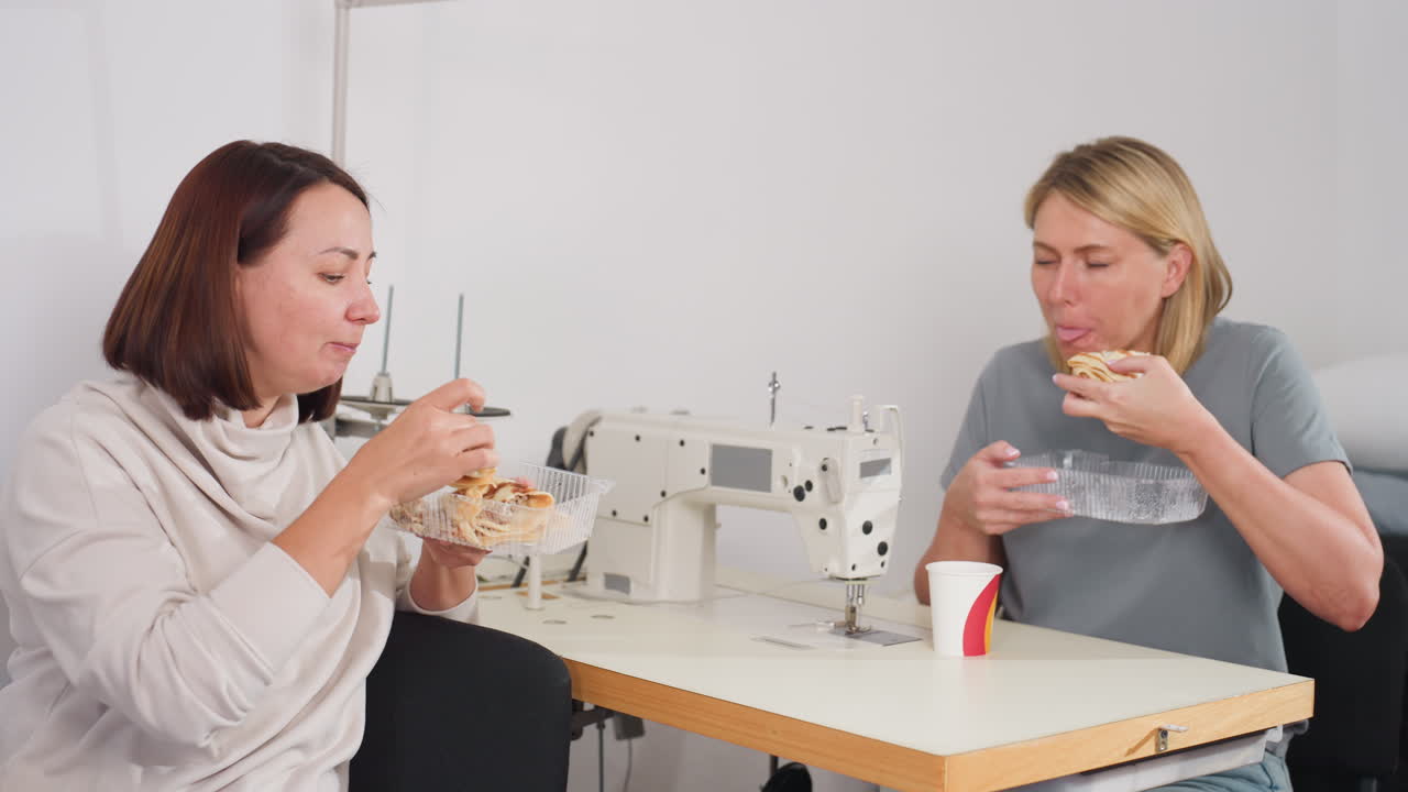 Two women seated beside sewing machines in bright organized tailoring workspace enjoying dessert together, eating happily during break time, smiling and sharing lighthearted moment