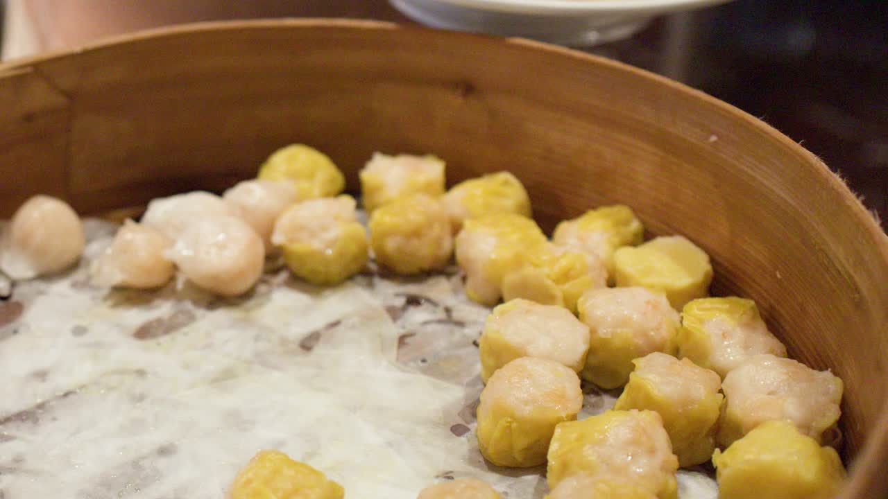 Primo piano di pinze metalliche che selezionano ravioli shumai gialli da un tradizionale cestello per la cottura a vapore in bambù.