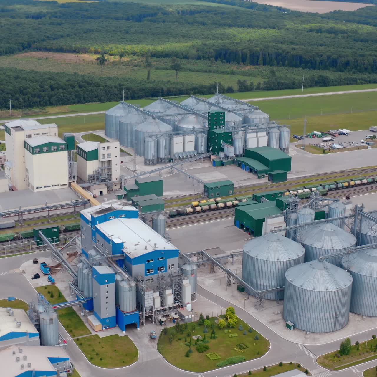 Large industrial complex in the countryside. Modern granary. Metal silos on field near beautiful green landscape. Aerial view. Camera rising