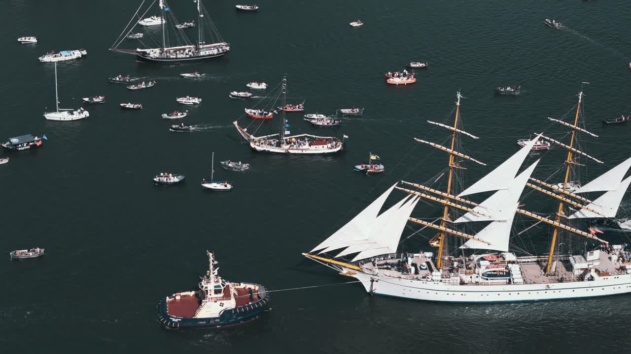 Tug boat tows white sailboat through crowd during Amsterdam Sail event, aerial medium static