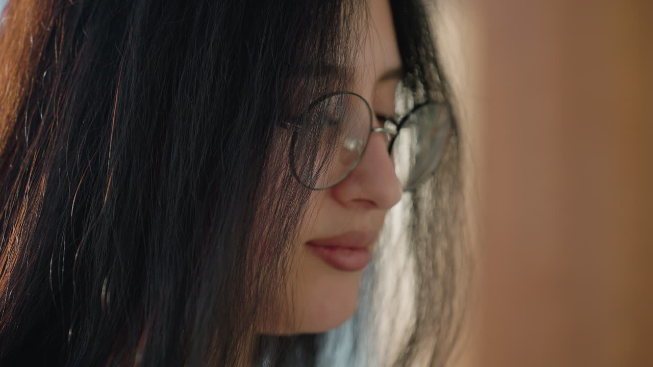 Charming close up of smiling lady wearing round glasses with long dark hair, softly lit by natural daylight in modern interior, captured in warm, serene moment of joy and reflection