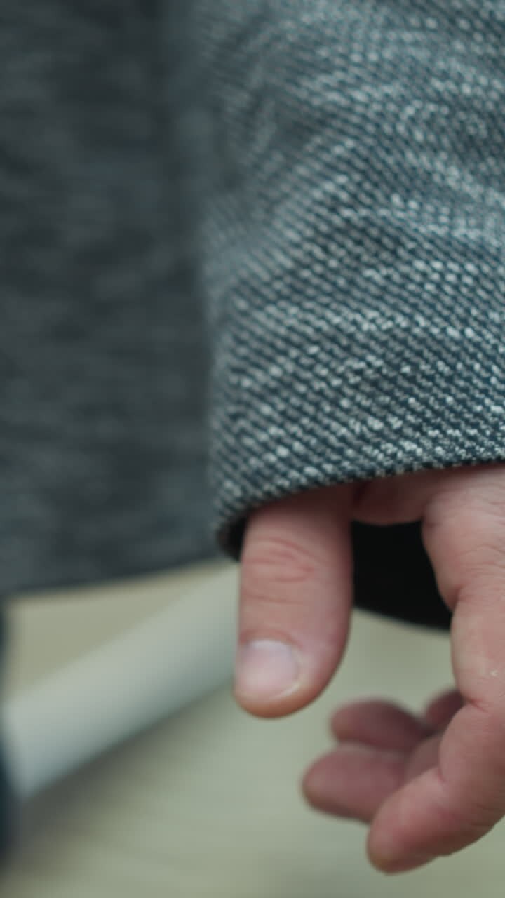 A close-up view of a man's hand and grey suit jacket sleeve, walking on a railway track, the surrounding is blurred, his attire includes a grey blazer and blue jeans