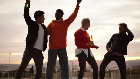 amigos multiétnicos bailando y bebiendo en un techo al caer la noche