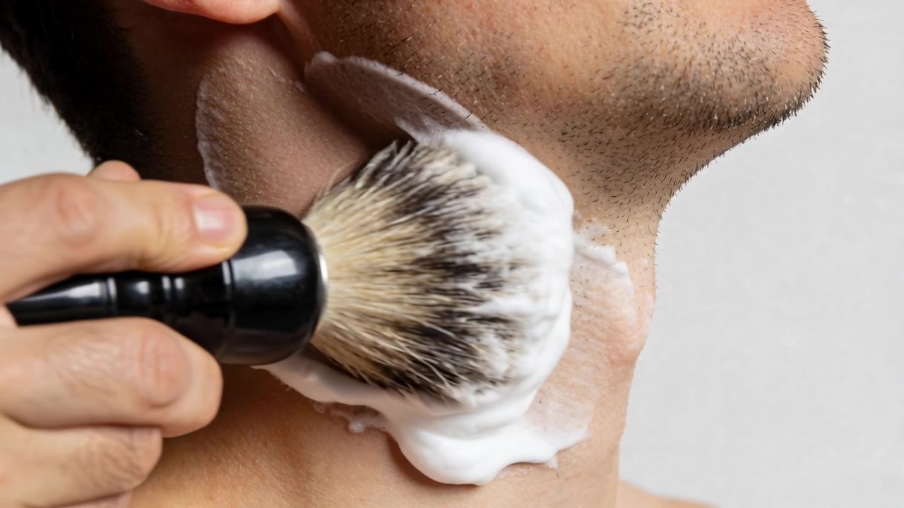 In primo piano, un pennello premuto contro il collo di un uomo, una mano che spalma una spessa schiuma da barba, preparandosi alla rasatura.