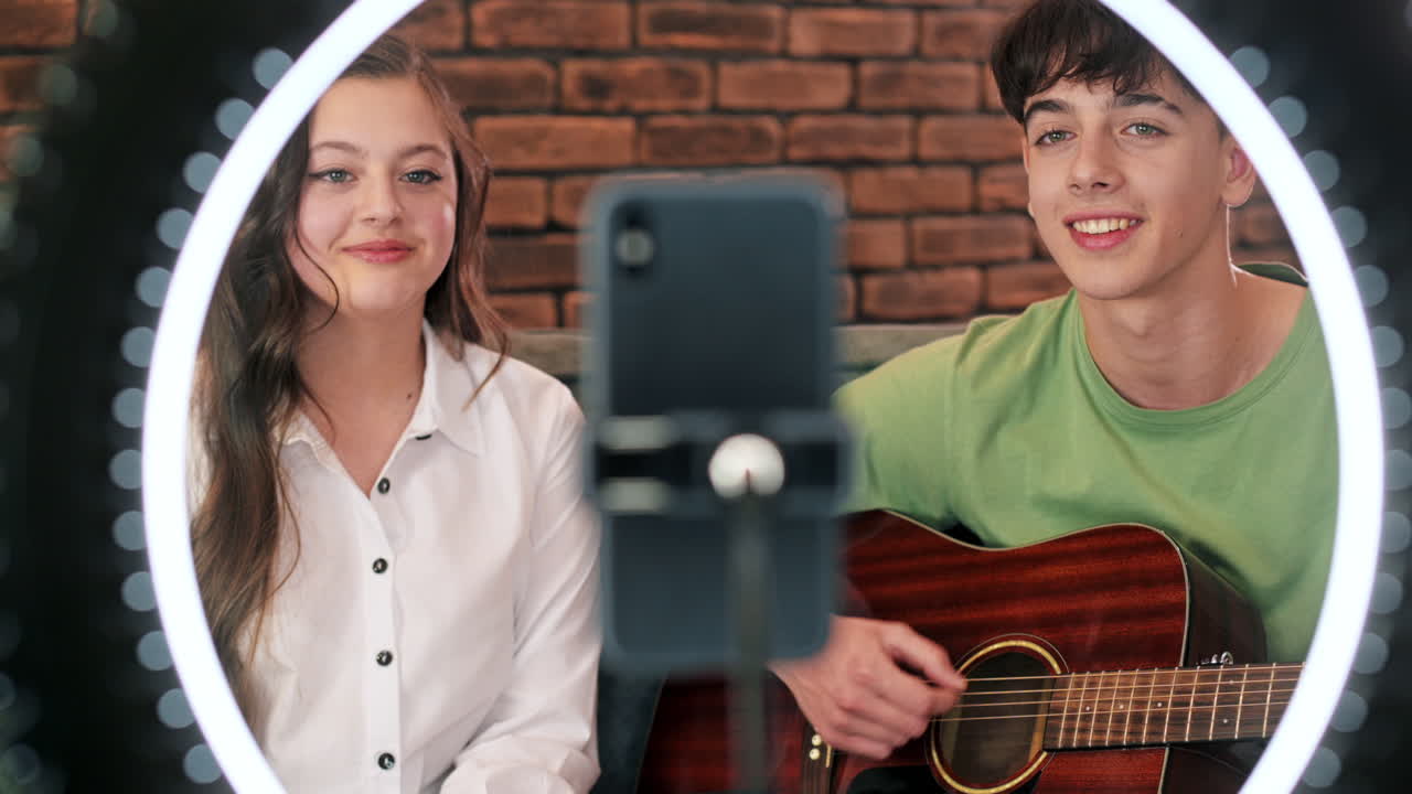 Ung man och kvinna som spelar gitarr och sjunger, filmar sig själva med en smartphone på ett stativ med ring light i en studio