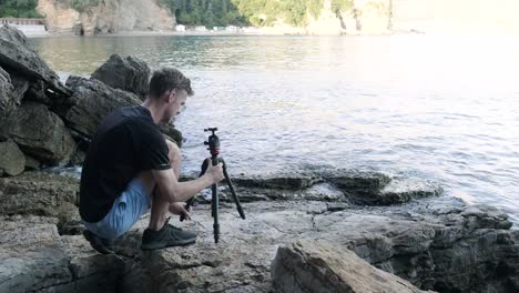 Ein-Mann,-Der-Seine-Kamera-Aufstellt,-Um-Die-Altstadt-Von-Budva-In-Montenegro-Mit-Wellen-Zu-Fotografieren,-Die-Während-Des-Sonnenuntergangs-Vor-Ihm-Zusammenbrechen