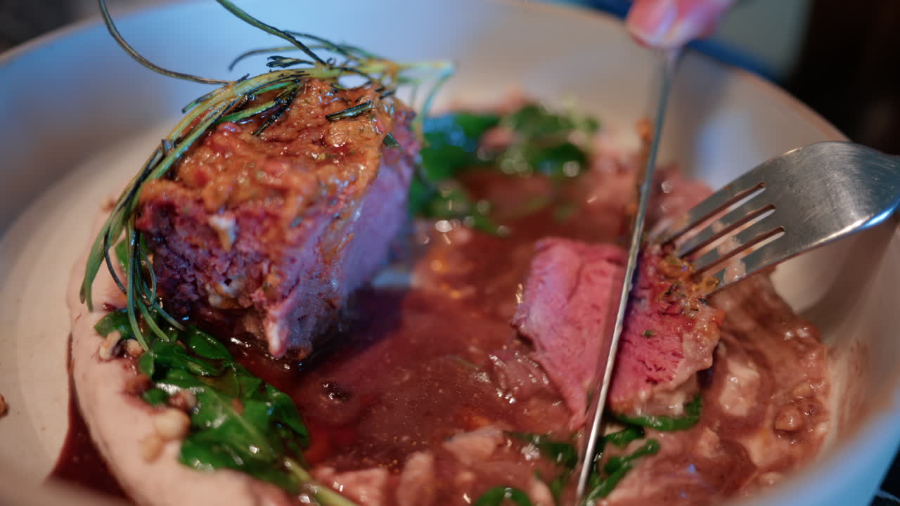 Close up of a woman's hand cutting into a gourmet meat dish served with sauce, greens, and rosemary garnish on a white plate