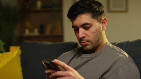 young man spending evening at home sitting on sofa with mobile phone scrolling through internet or social media streaming content 1