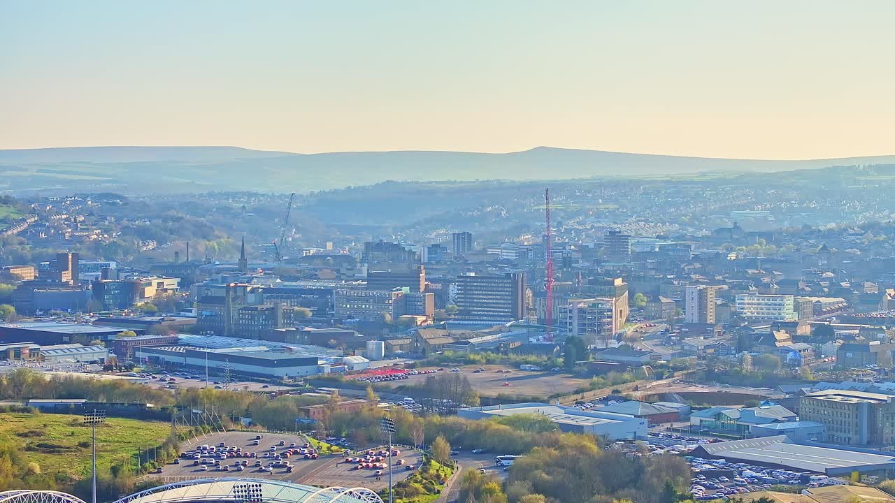 Elevated drone footage showing the Huddersfield Town Football Club area and cityscape, featuring commercial buildings, stadium outlines, cranes, and rolling hills in warm morning light