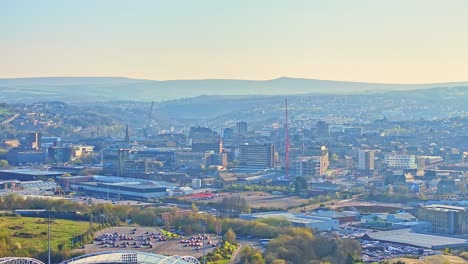elevated drone footage showing the huddersfield town football club area and cityscape, featuring commercial buildings, stadium outlines, cranes, and rolling hills in warm morning light