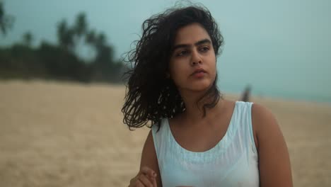 A-woman-in-a-white-sleeveless-top-stands-on-a-sandy-beach-with-her-curly-hair-blowing-in-the-wind