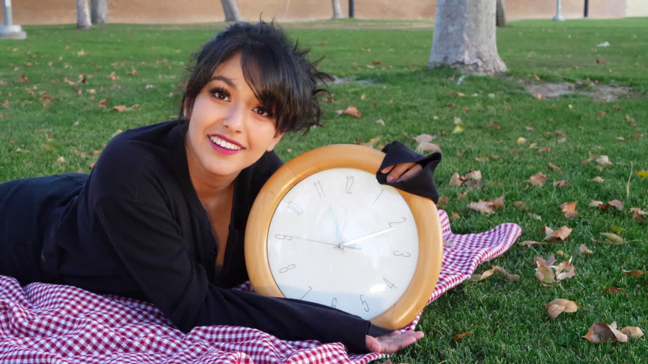 una chica bastante atractiva sonriendo con un gran reloj para mostrar el tiempo en un parque durante el otoño con hojas en el suelo y espacio para copiar