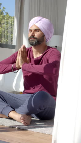 vertical video: meditating at home, indian man in turban sitting cross-legged with hands in prayer