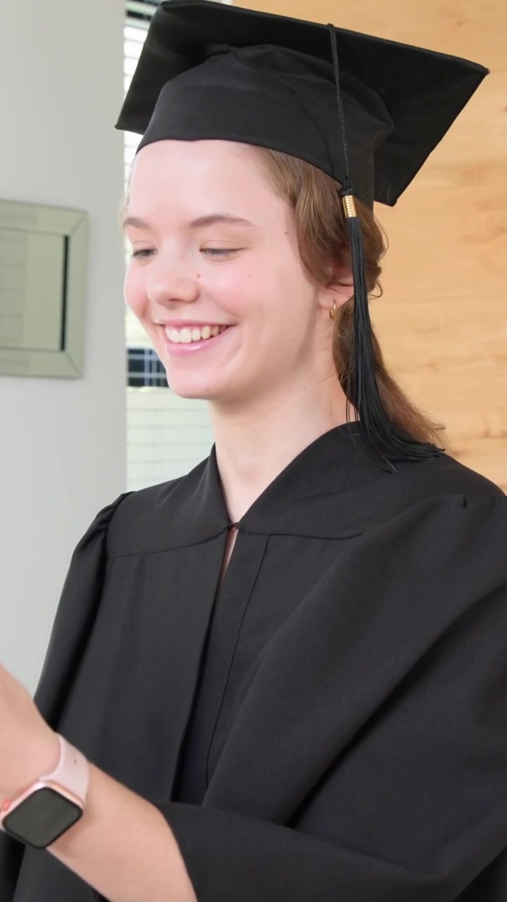 Vertical video: Waving, graduate wearing cap and gown tapping phone at home, checking messages