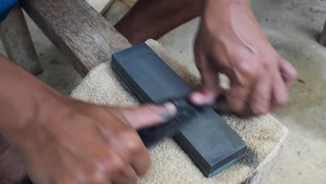 man sharpening a machete with a whetstone on a small wooden bench