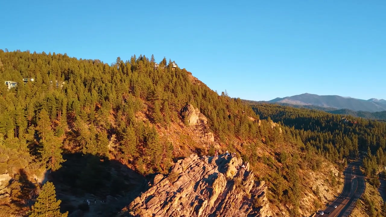 Cresta rocciosa di una grotta sotto pendii di conifere nella luce serale. La luce arancione del sole tocca un affioramento frastagliato e gli alberi, con una strada e montagne in lontananza sulla destra
