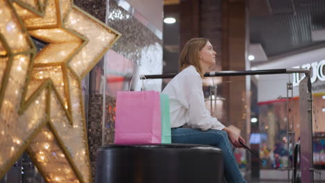 lady seated on bench in shopping mall holding tablet surrounded by festive decorations, illuminated star decor, blinking lights, and shopping bags, with escalators and lively background