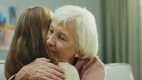 portrait of the senior smiled caucasian gray haired woman hugging her little granddaugter while they sitting on the sofa at home. rear. close up. indoors.