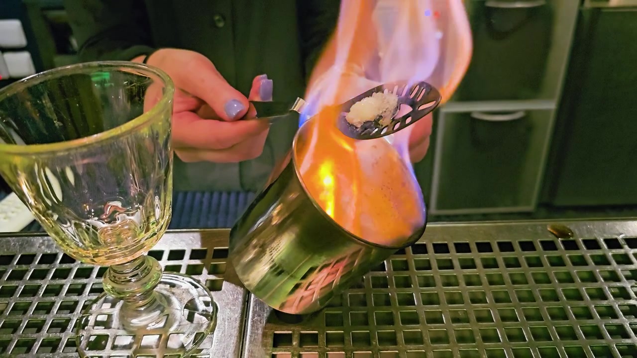 Vista dettagliata del bancone di un bar con un barista che prepara un cocktail infuocato, con fiamma blu, zolletta di zucchero, cucchiaio forato, tazza di metallo, calice vuoto, riflessi verdi e atmosfera da night club.