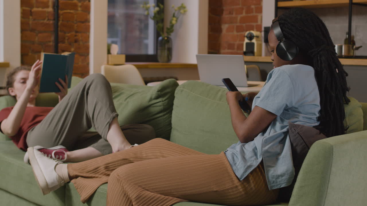 집에서 소파에서 스마트폰으로 책을 읽고 음악을 듣는 소녀들