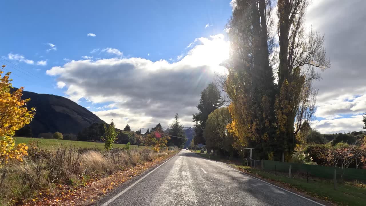 Vehicle drives along sunlit rural road lined with autumn trees, mountains, and open fields
