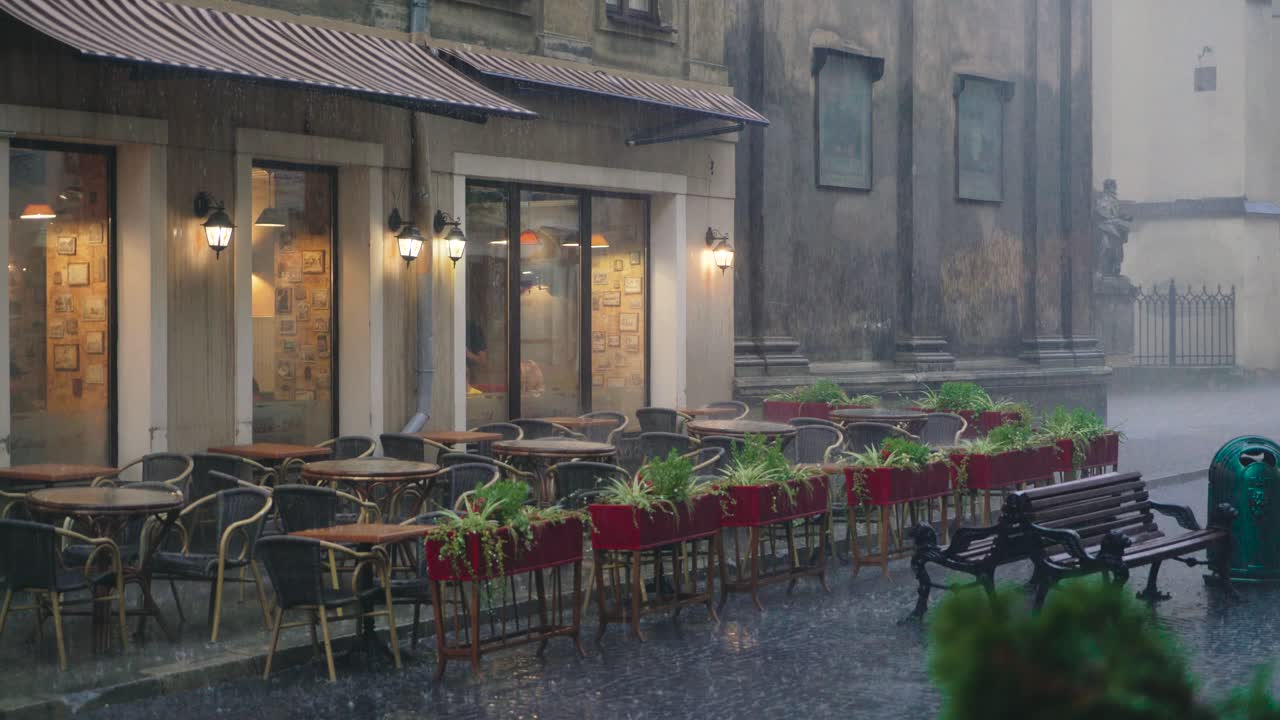 raindrops are falling on the tables and chairs of the restaurant on the street. Wet summer terrace with flower pots is near the road