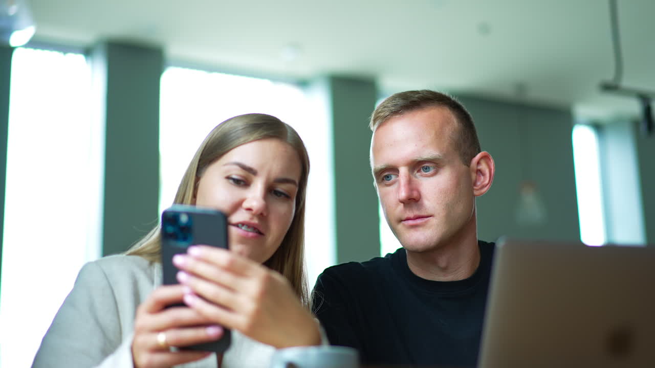 Two mid-aged people sitting indoors look at smartphone in lady's hands. Couple talks and discusses something on the phone. Blurred backdrop.