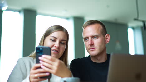 two mid-aged people sitting indoors look at smartphone in lady's hands. couple talks and discusses something on the phone. blurred backdrop.