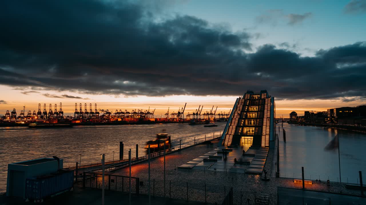 Passerande moln och fartyg under solnedgången i Hamburgs hamn och containerterminal vid floden Elbe med upplysta lampor på en modern glasbyggnad