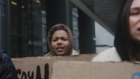 Young-American-Female-Activist-Holding-A-Cardboard-Placard-During-A-Climate-Change-Protest-While-Looking-At-Camera-Surrounded-By-Others-Activists