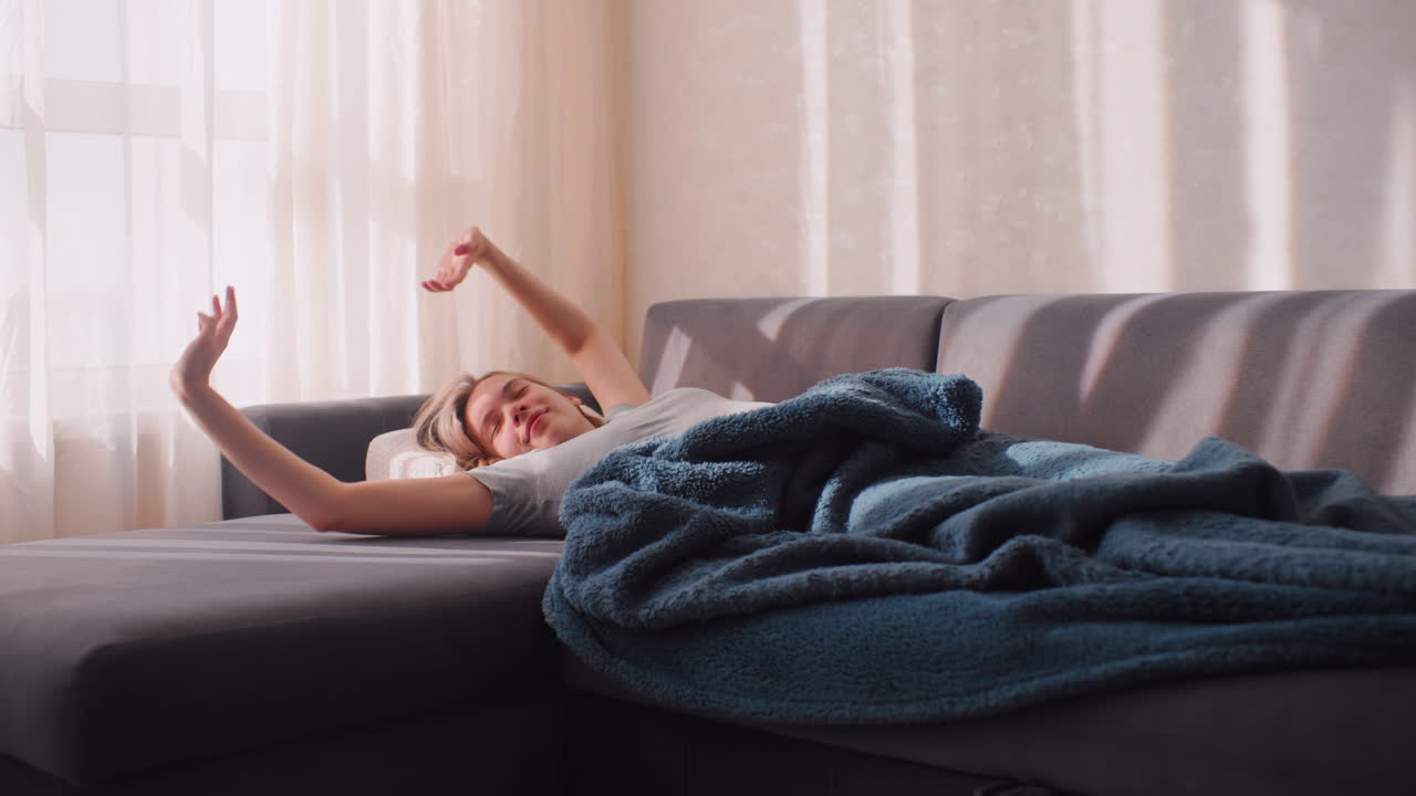 Boss lady unwinds with warm smile as she stretches after sleep on sofa in bright living room, resting under cozy blanket with soft morning sunlight shining through curtains