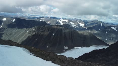 aerial drone fotoage flying over tall dark black colored kebnekaise mountain tops that have white snow patches between them while the sun is shining cloud shadows are visible between mountains, hills.