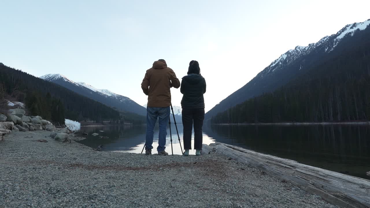 Coppia che fotografa la calma superficie del lago Duffey che riflette montagne innevate e foreste di conifere nella Columbia Britannica, Canada