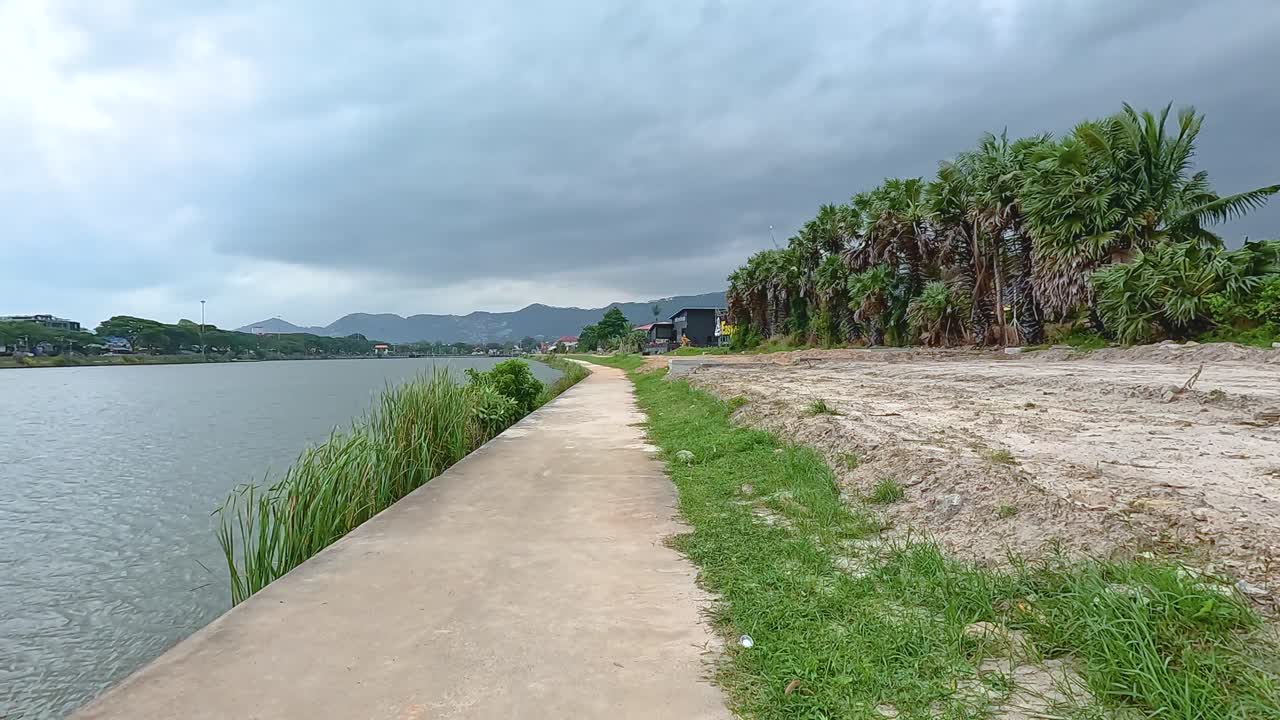 A tranquil riverside path in Koh Samui, bordered by lush greenery and calm waters under overcast skies
