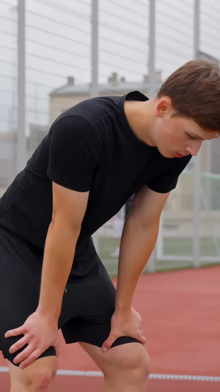 Young man in a black t-shirt leans forward to catch his breath after running, standing on outdoor urban track as he recovers energy and strength during solo fitness training session, vertical reveal