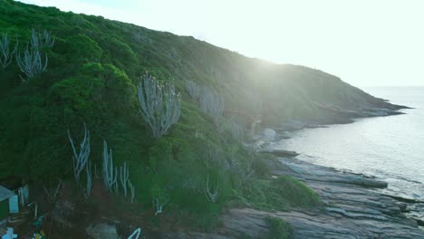 órbita-Aérea-Nostálgica-De-Un-Rayo-De-Sol-A-Través-De-Una-Montaña-Con-Vegetación-Tropical-Y-Rocas-Con-Olas-Suaves,-Costa-De-Playa-Joao-Fernandes