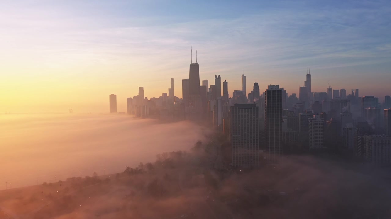 Smooth aerial approach toward the Chicago skyline as dense morning fog glows orange and gold along the lakefront beneath the rising sun.