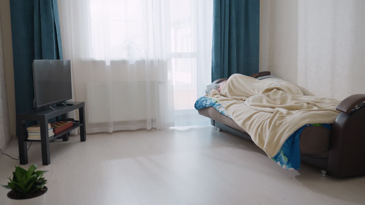 Individual sleeping turning on bed under blanket in modern bright bedroom interior with sunlight streaming through window covered by white curtain next to blue drape