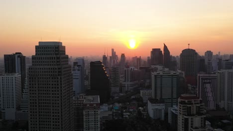 epic aerial flying through buildings, skyline and skyscrapers of sukhumvit district during colorful red sunrise in bangkok, thailand.