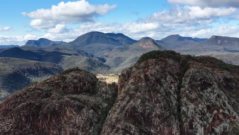 drone camera glides above rugged split rock, revealing expansive mountain ranges and valleys under bright daylight with scattered clouds in rural australia