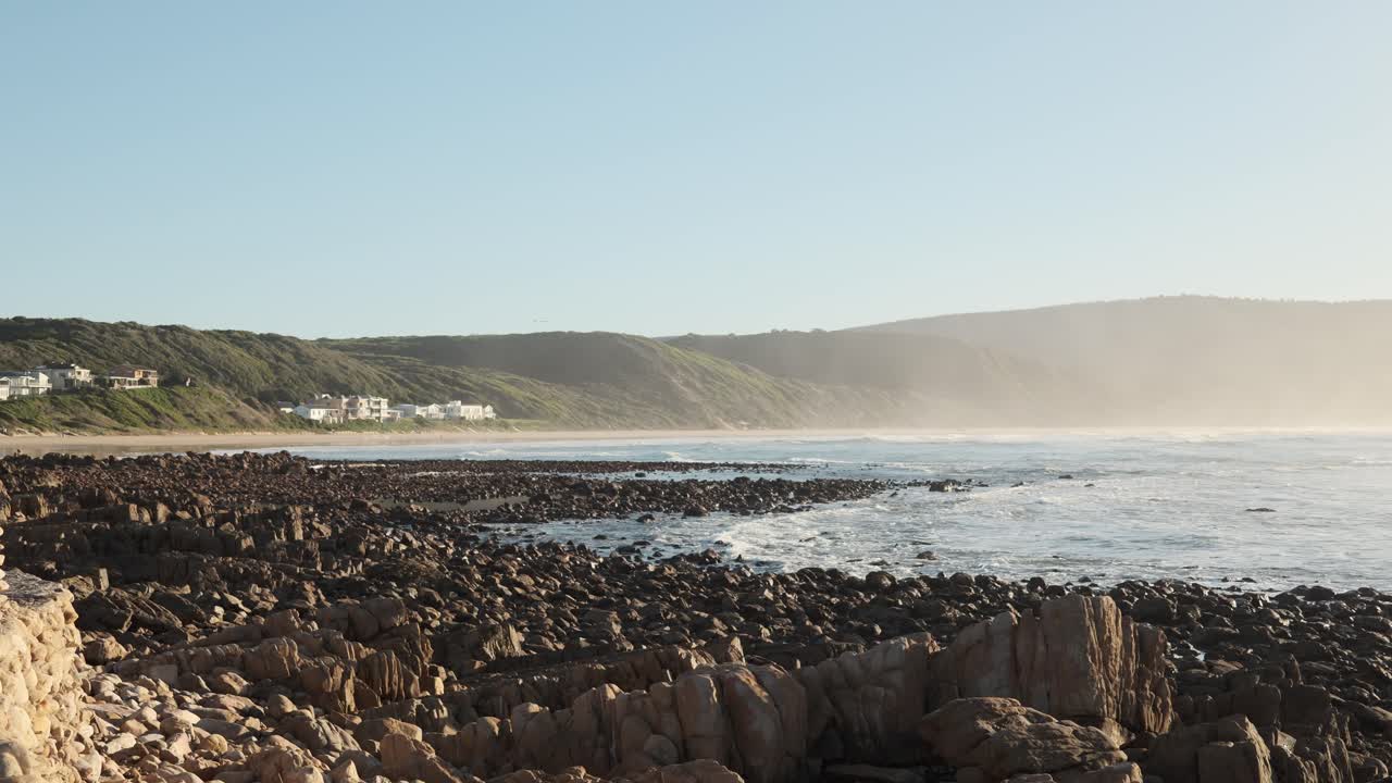 Static 4K shot of a low tide sea revealing many rocks. Mist covers mountains in the background under a sunny, warm day in South Africa’s Western Cape. A peaceful coastal landscape