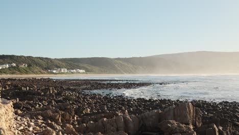 static 4k shot of a low tide sea revealing many rocks. mist covers mountains in the background under a sunny, warm day in south africa’s western cape. a peaceful coastal landscape
