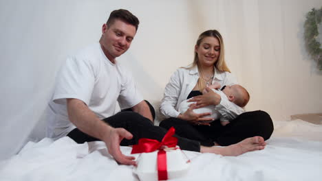 parents and their child sit on the bed. mom waving her baby and dad takes a box with red ribbon.