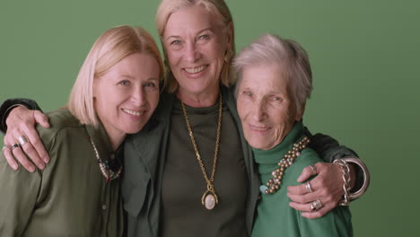 two blonde mature women and blonde senior woman smiling and hugging, wearing green tones clothes and posing on green background