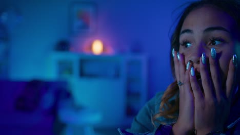 close up portrait of a beautiful excited young black girl watching an action video on a computer. she is mind blown. screen adds reflections to her face. cozy room is lit with warm light.