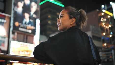 Woman-in-black-stands-on-a-bridge,-looking-over-a-lively-cityscape-at-night